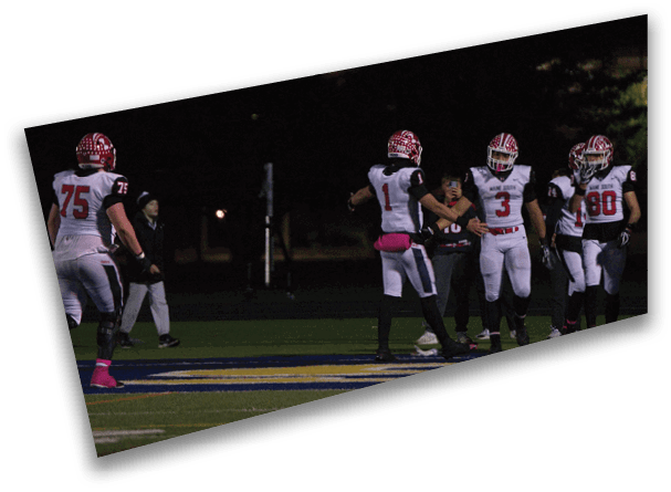 People wearing football uniforms are standing on a field at night, with some interacting and one person holding a phone. Description generated by AI