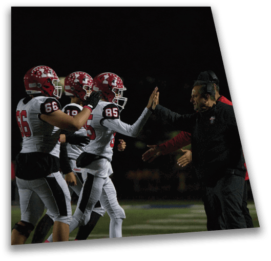 People in football uniforms are giving high fives to coaches on the sidelines during a nighttime game. Description generated by AI