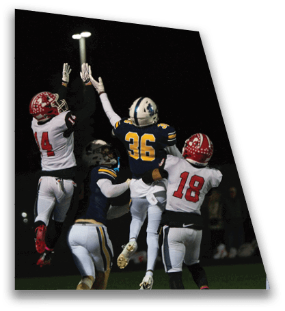 People in football uniforms are jumping and reaching up to catch or block a ball during a nighttime game under bright stadium lights. Description generated by AI