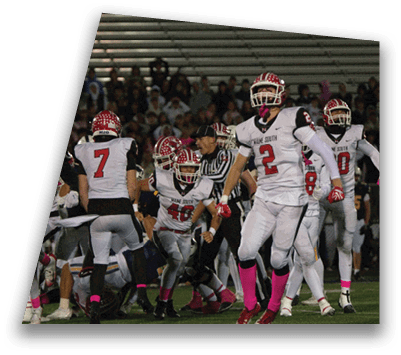 Several football players in white uniforms with red helmets and pink accessories are actively engaged on a field with a referee nearby and spectators in the background. Description generated by AI