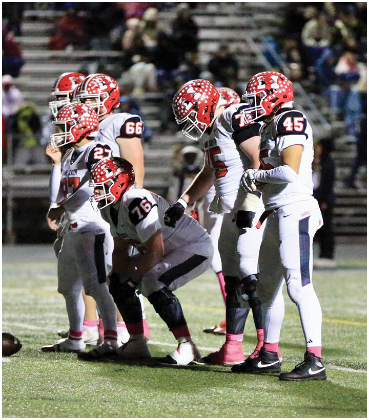Several people in football uniforms and helmets are positioned on a field, appearing to prepare for a play during a game with spectators in the background. Description generated by AI