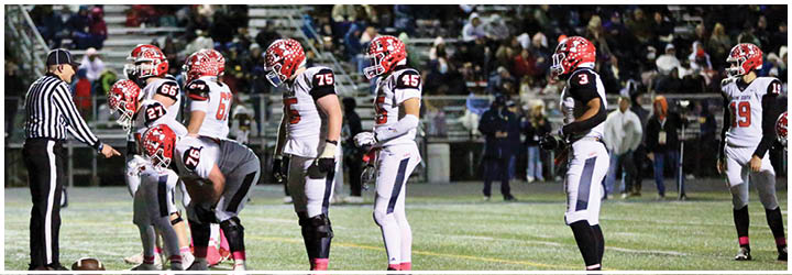 A group of football players in helmets and uniforms are lined up on a field with a referee standing nearby, preparing for a play. Description generated by AI