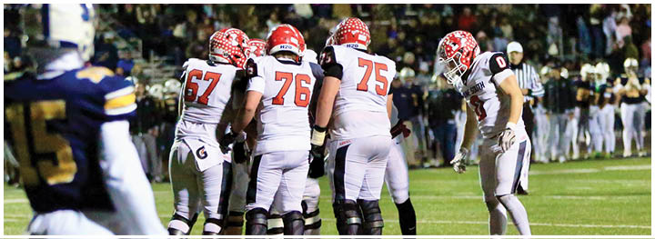 People in football uniforms are gathered on a field during a nighttime game with spectators in the background. Description generated by AI