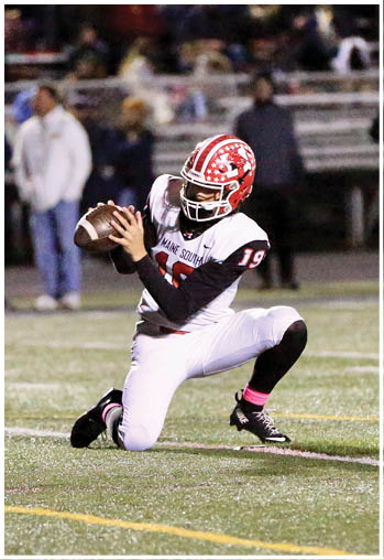 A person wearing a red and white football helmet and uniform is kneeling on one knee on a football field while holding a football. Description generated by AI