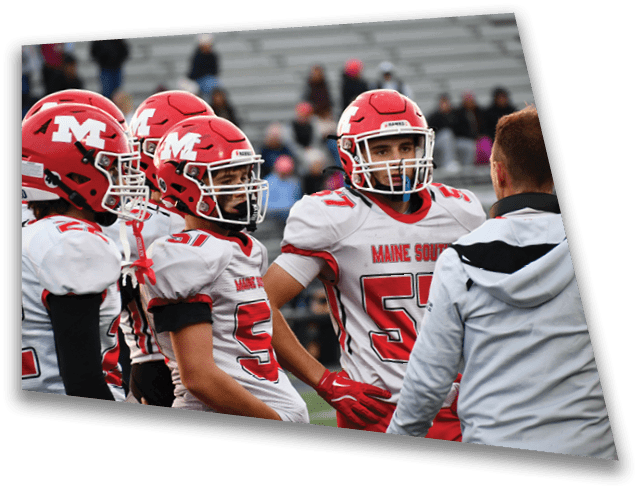 Two people in red and white football uniforms with helmets are standing on a field, engaged with a person in a gray and black jacket. Description generated by AI
