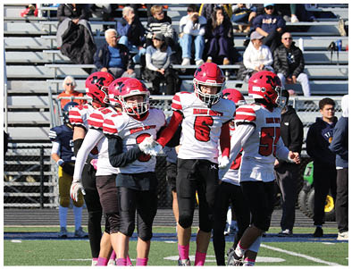 People in football uniforms and helmets stand on a field near two adults who are conversing by the sidelines with empty bleachers in the background. Description generated by AI