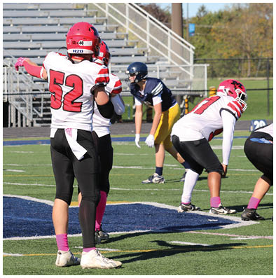 People wearing football uniforms are positioned on a field with bleachers in the background, with one person in a white jersey numbered 52 standing with arms outstretched. Description generated by AI