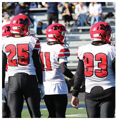 Two people wearing red helmets and white sports jerseys with large red numbers are standing close together on a sports field. Description generated by AI