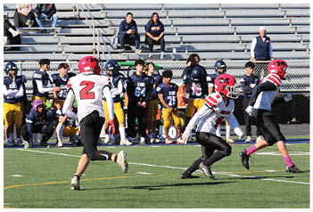 Several people in sports uniforms are playing football on a field with a mostly empty bleacher in the background and a sign that reads “BLUE CREW." Description generated by AI