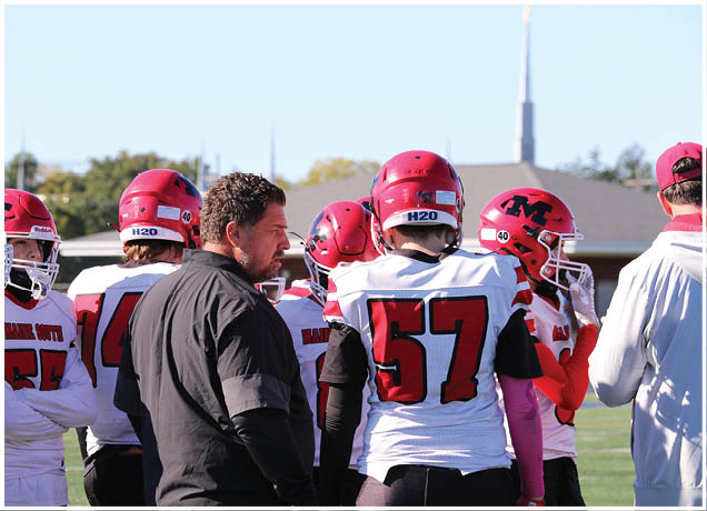 A group of people wearing red and white football uniforms and helmets are gathered outdoors with one person in a black jacket appearing to give instructions. Description generated by AI