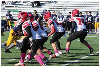 A group of people wearing football helmets and uniforms are actively engaged in a play on a football field with empty bleachers in the background. Description generated by AI