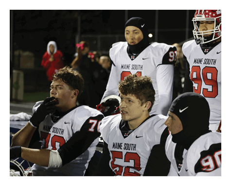 Five people wearing white football uniforms with “Maine South" on the front are sitting and standing together on a sports field at night. Description generated by AI