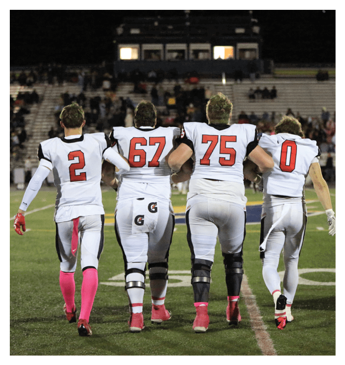 Four people in white football uniforms with red numbers walk arm in arm on a field toward a stadium with spectators in the background. Description generated by AI