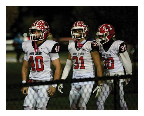 Three people wearing white football uniforms with red helmets and the words “Maine South" walk together on a field at night. Description generated by AI