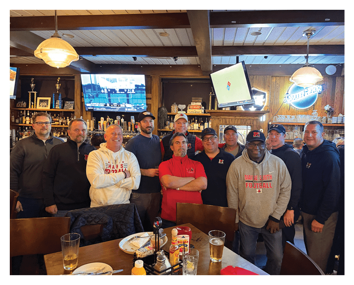 A group of nine adults wearing casual clothing and sports-themed apparel stand closely together inside a bar with wooden walls, televisions, and various decorations in the background. Description generated by AI