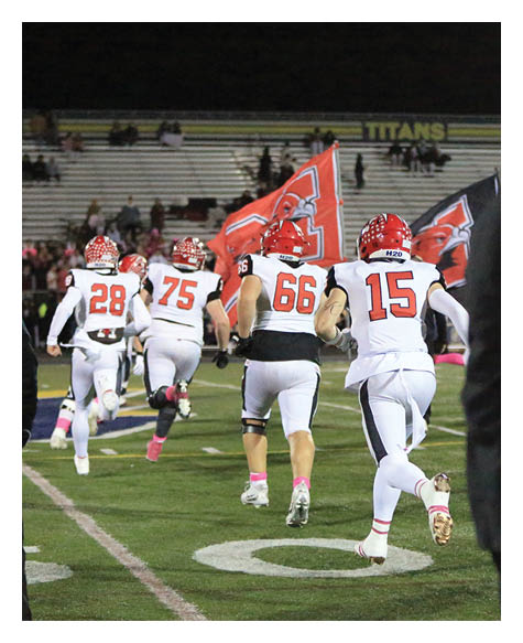Several people in football uniforms with red helmets are running on a field toward a crowd holding red flags, while a person in dark clothing and a cap stands in the foreground. Description generated by AI