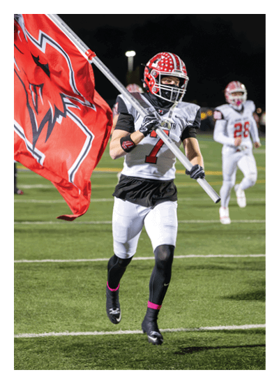A person in a football uniform is running on a field holding a large flag while another person in a similar uniform runs in the background. Description generated by AI