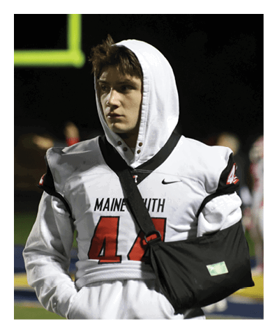 A person wearing a white football jersey with the number 44 and a white hooded sweatshirt underneath stands on a field at night with one arm in a black sling. Description generated by AI