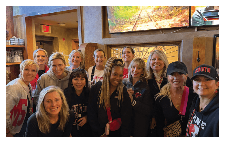 A group of smiling adults posing closely together inside a warmly lit restaurant with wooden decor and colorful artwork on the walls. Description generated by AI
