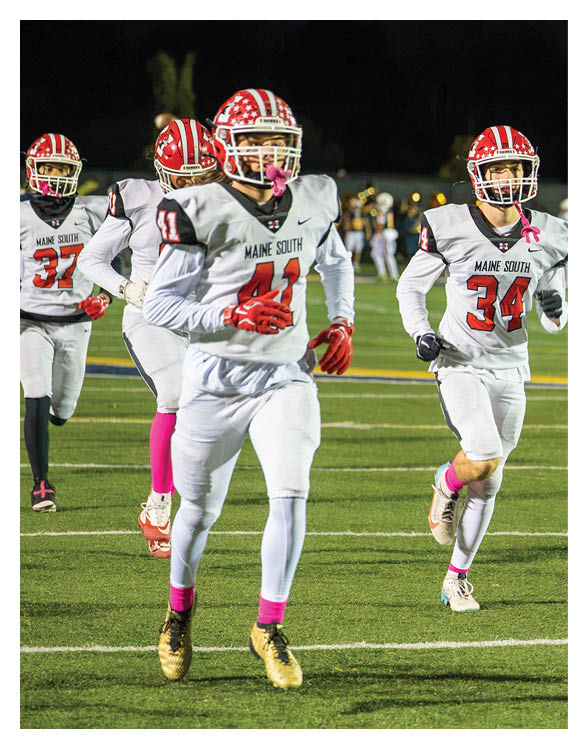 Two people wearing football helmets and white jerseys with “Maine South" on the front are on a field at night. Description generated by AI