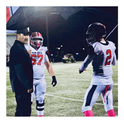 Three people in football uniforms stand on a field, with one person wearing a leg brace and gray sweatpants while the others wear full gear including helmets and pink socks. Description generated by AI