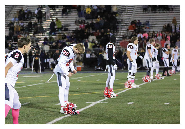 Three people in football uniforms stand on a field with two helmets on the ground, one person looking down at a helmet and another wearing bright pink socks. Description generated by AI