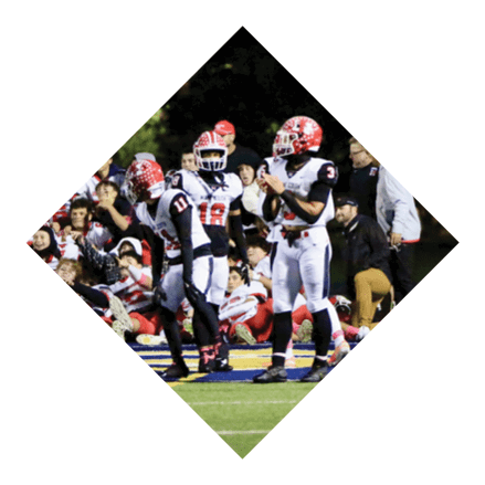 A group of people in football uniforms are on a field with some sitting on the sidelines and others standing, appearing to be engaged in a game or practice at night. Description generated by AI