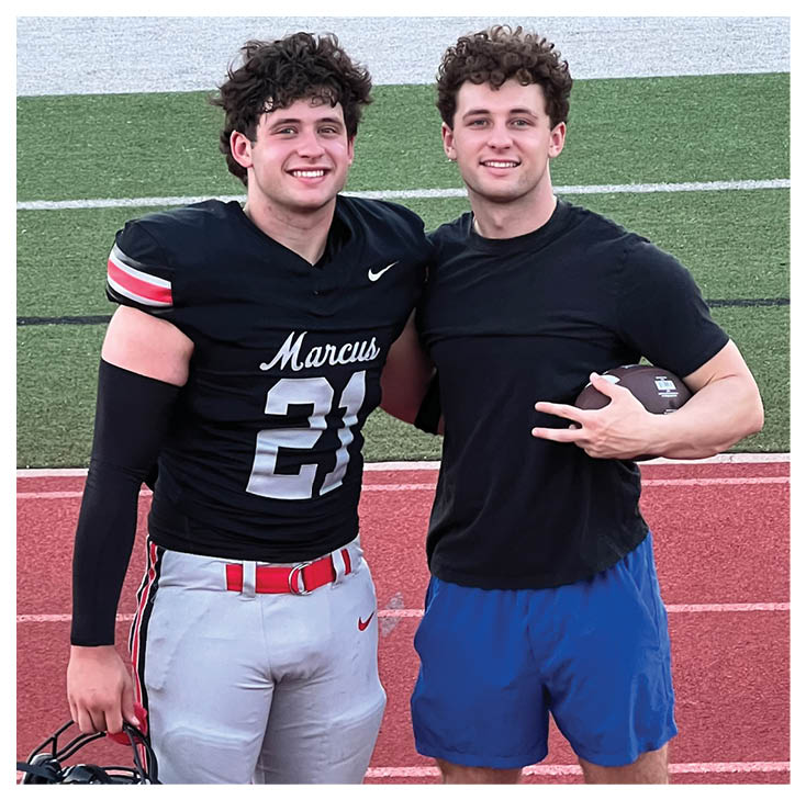 Two people stand side by side on a red running track, one wearing football pants and cleats while holding a helmet, and the other wearing blue shorts and casual slip-on shoes. Description generated by AI