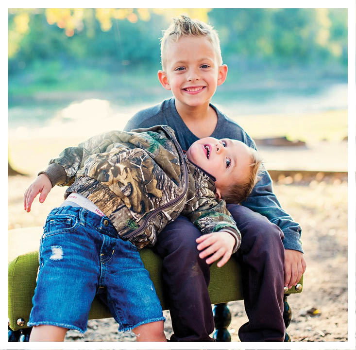 Two children are outdoors with one child smiling and holding the other child who is wearing a camouflage jacket and looking up. Description generated by AI