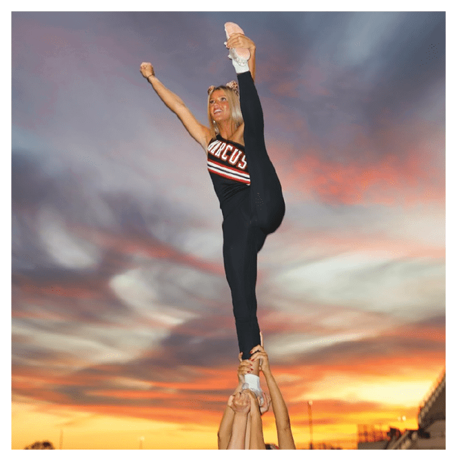 A person wearing a cheerleading outfit is performing a high leg stretch with one arm raised against a colorful evening sky. Description generated by AI