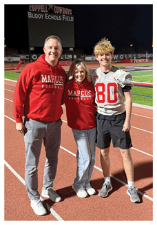 Three people stand on a running track at night with a football field and scoreboard in the background, two wearing matching red hoodies with “Marcus Football" and one wearing a football jersey with the number 80. Description generated by AI