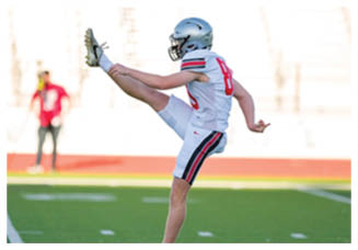 A person in a football uniform and helmet is kicking a football on a field with a blurred background. Description generated by AI