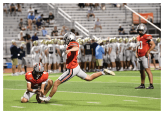 Three people in red and gray football uniforms are on a field with one person holding the ball on the ground while another kicks it, and a third stands nearby with a blurred crowd in the background. Description generated by AI