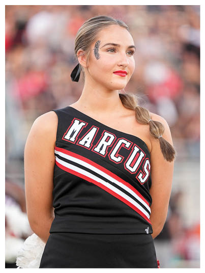 A person with braided hair wearing a black one-shoulder cheerleading uniform with the word Marcus and red, white, and black stripes stands with a confident expression and glitter decoration near one eye. Description generated by AI