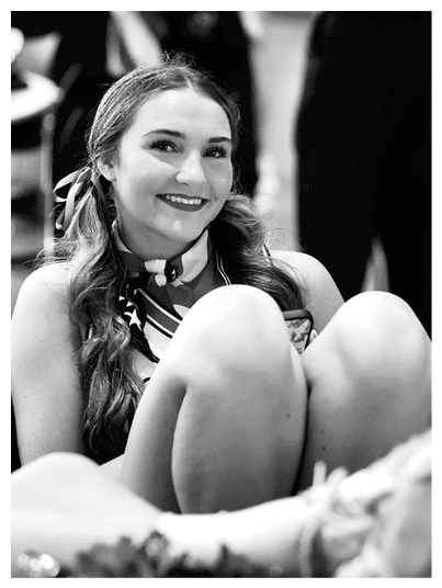 A person with long wavy hair tied with a ribbon is smiling and wearing a scarf around their neck while sitting with one knee raised. Description generated by AI