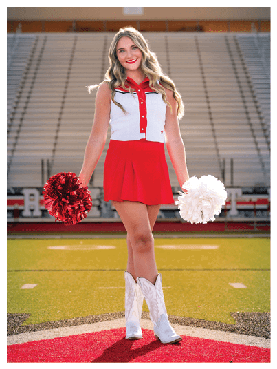 A person with long wavy hair wearing a red and white cheerleading outfit and white boots is standing on a football field holding red and white pom-poms with empty bleachers in the background. Description generated by AI