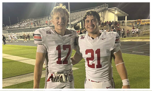 Two people wearing white football uniforms with numbers 17 and 21 stand on a field at night smiling with their arms around each other. Description generated by AI