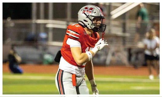 A person wearing a red football jersey and helmet is standing on a football field, appearing to be in a ready or focused stance. Description generated by AI