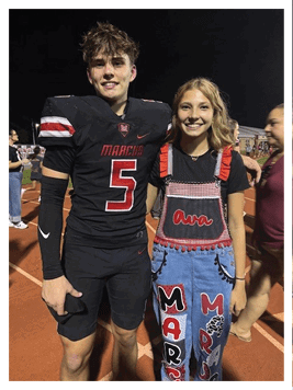 Two smiling people stand close together on a sports field at night, one wearing a sports jersey and the other wearing a black shirt with red and white checkered overalls embroidered with the name Ava. Description generated by AI