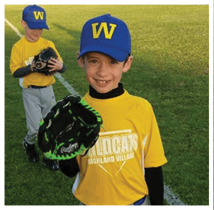 A child wearing a blue cap with a yellow “W" and a yellow Wildcats Highland Village baseball jersey is smiling and holding a black and green baseball glove on a grassy field. Description generated by AI
