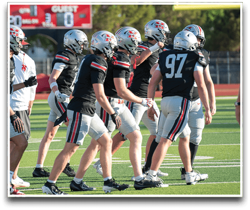 A group of people wearing football helmets and black jerseys with white pants are walking on a green football field near a scoreboard. Description generated by AI