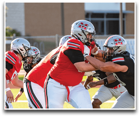 Several people wearing football helmets and uniforms are engaged in a blocking action on a football field during a game. Description generated by AI