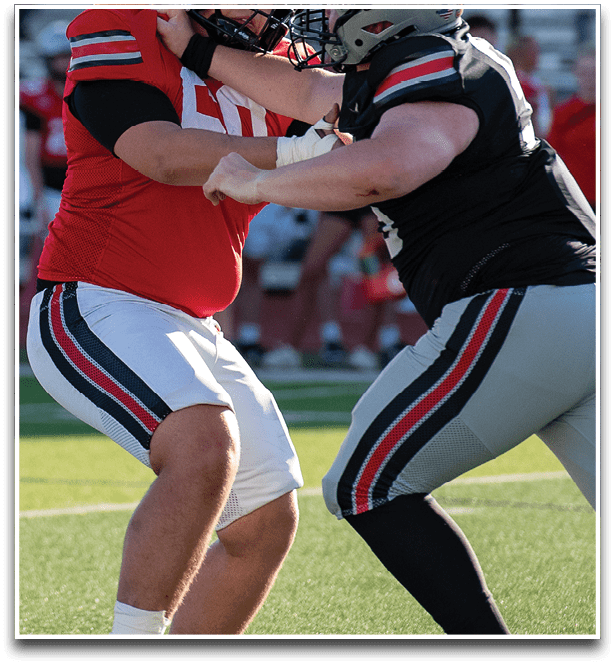 Two people wearing football uniforms are engaged in a blocking stance on a grassy field during a game or practice. Description generated by AI