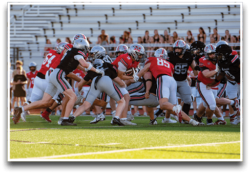 A group of football players in red and black uniforms are engaged in a play on a green field with empty bleachers in the background. Description generated by AI