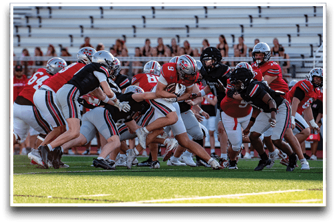 A group of people wearing football uniforms are engaged in a play on a football field with empty bleachers in the background. Description generated by AI