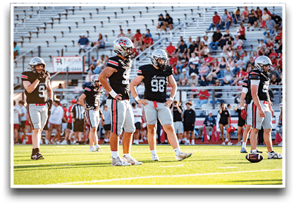 Several people wearing black football uniforms and helmets stand on a green field with a crowd seated in bleachers in the background. Description generated by AI
