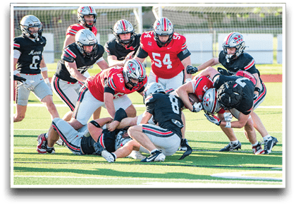 Several people wearing football helmets and uniforms are engaged in a tackle on a grassy field during a game. Description generated by AI