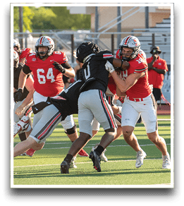 Two people in red football uniforms are blocking two people in black football uniforms during a game on a sunny field. Description generated by AI