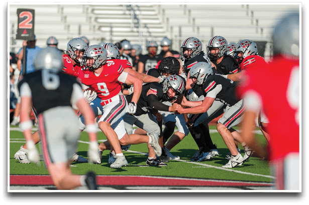 Players in red and black football uniforms are engaged in a play on a green field with a 2nd down marker visible in the background. Description generated by AI