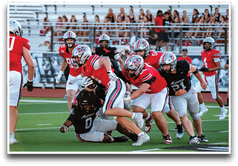 Several people in red and black football uniforms are engaged in a tackle on a green field with a crowd in the background. Description generated by AI